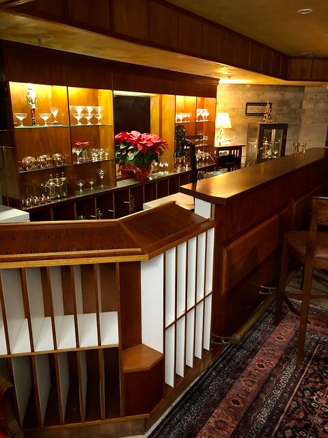 Speakeasy wood-paneled bar with wine glasses in lighted cabinet above the bar.