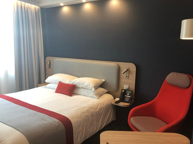 Holiday Inn Express Paris double hotel room with gray walls, white pillows, white sheets and red accent pillow. Red chair and laminate table are next to bed. 