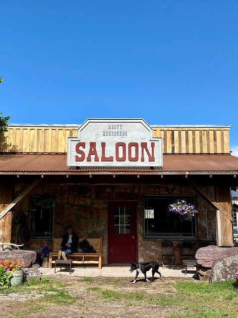 A cowboy relaxes on a wooden bench in front of the Rusty Horseshoe Saloon, while a black dog walks by. 