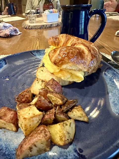 Breakfast croissant sandwich with scrambled egg, cheese, bacon and roasted, diced potatoes served on a blue ceramic plate. A blue ceramic coffee mug is beyond the plate. 