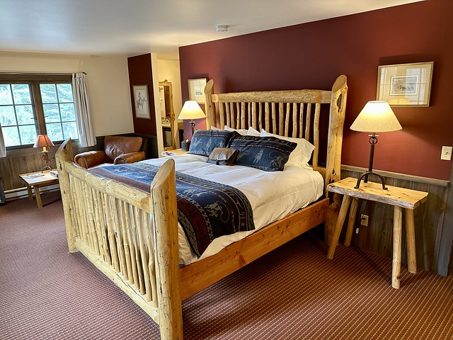 Light wood king bed frame with white duvet and western-themed runner and 2 decorative pillowcases. Matching wood side table and desk lamp on either side of bed. A large light brown leather chair is to left of bed with cowboy on horse artwork above the chair. The wall is painted red. Brick red carpet with beige strips is on 2nd-story lodge room. 