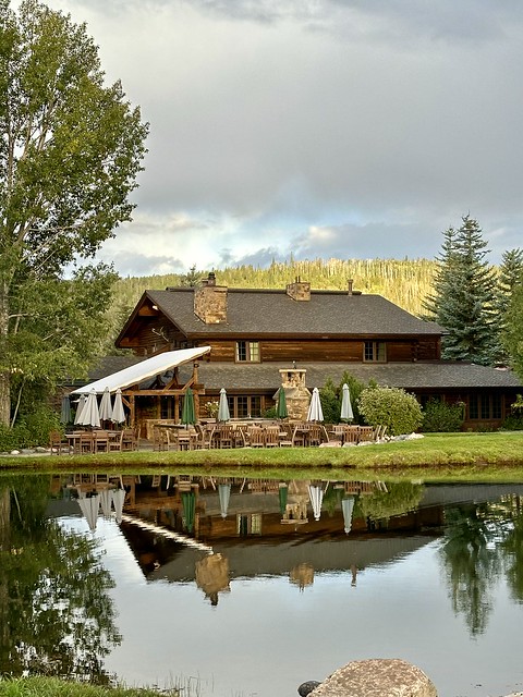 Two-story wood lodge and outdoor patio furniture and umbrellas reflected in the lake next to the lodge. 