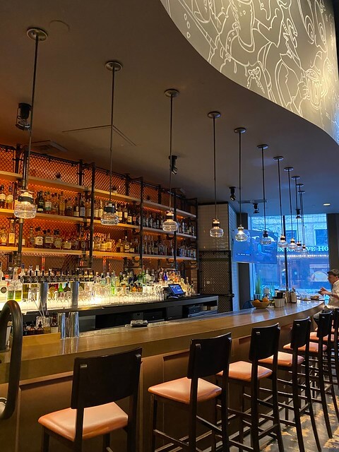 Outlier bar with wooden bar stools, suspended lighting and bottles of alcohol lighted for display.