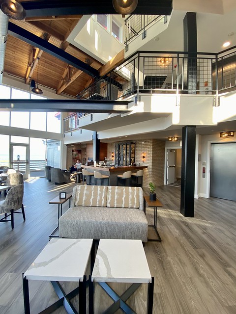 A three-story look into the interior of Cannery Pier Hotel & Spa. The ground floor features a small sofa and 2 white tables. Behind the furniture is Bar 600, the new hotel bar in Astoria. A wood, L-shaped counter surrounds 4 beige leather and dark metal bar stools. A small bar is located in the corner of the hotel lobby. 