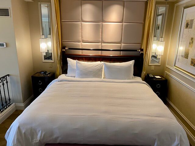 Oversized king bed with white bed cover and white pillows. Beige headboard and drop-down hanging light fixtures next to small wooden end tables on either side of bed. 