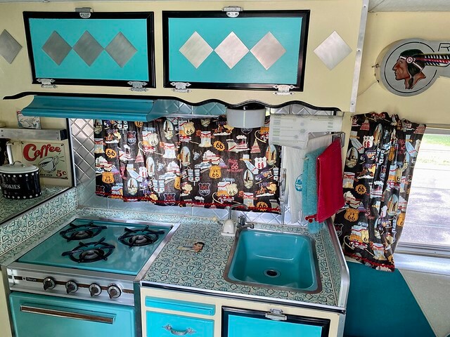 Inside the blue and white vintage trailer is a colorful kitchen filled with vintage kitchen utensils, a retro stovetop and sink. Route 66 motel themed curtains hang in the retro trailer. 