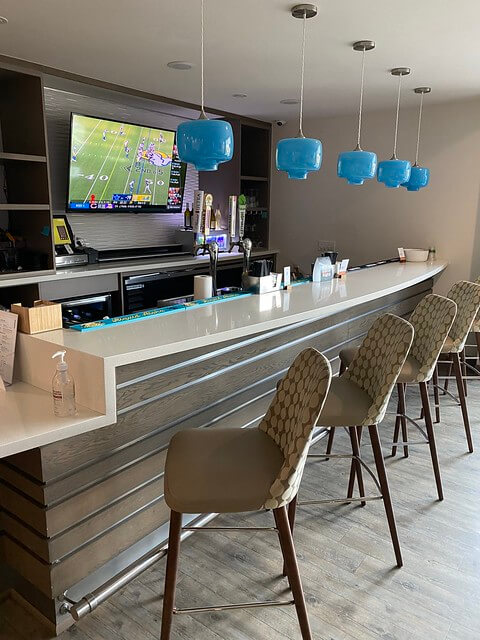 Even Bar with counter, blue handing light sconces and four bar chairs at the counter. A flatscreen tv is showing a University of Oregon football game. 