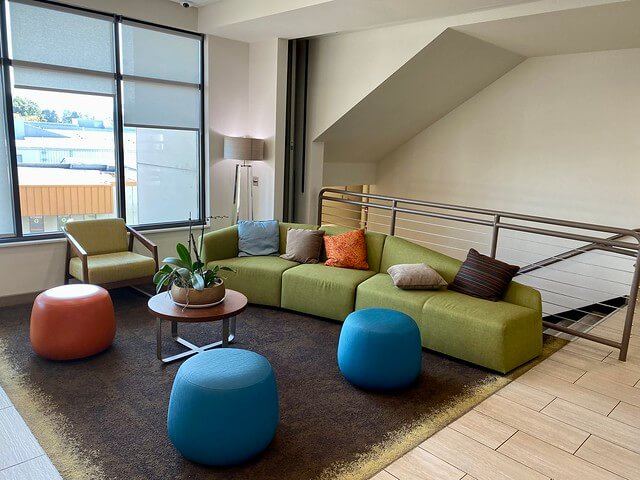 Long green couch with circular wood table and plant are available in the Even Hotel Eugene. Three soft, solid-colored stools are across from the sofa. 