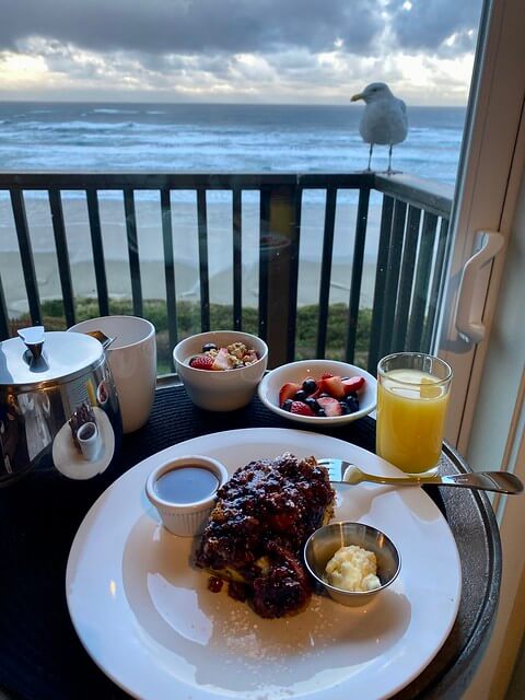 Marionberry French toast with syrup, coffee, fresh fruit plate and orange juice are served in the hotel room. Seagull perched on balcony railing, looking into hotel room. 