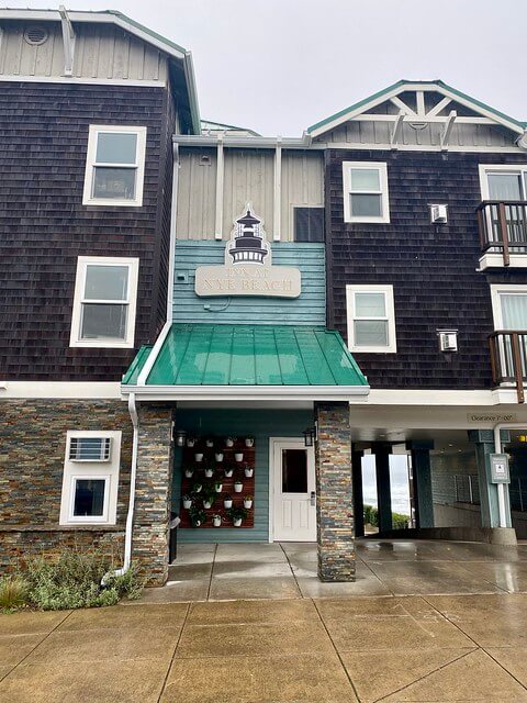 Inn at Nye Beach exterior with lighthouse logo sign above entrance. Stone and dark wood shingle exterior with hotel room windows face the street in Newport, Oregon. 