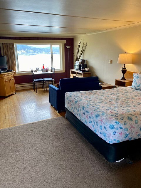 Queen bed with nautical themed bedspread, blue sofa and small table for two in front of large picture window. Window looks out to Depoe Bay and Pacific Ocean on Oregon coast.