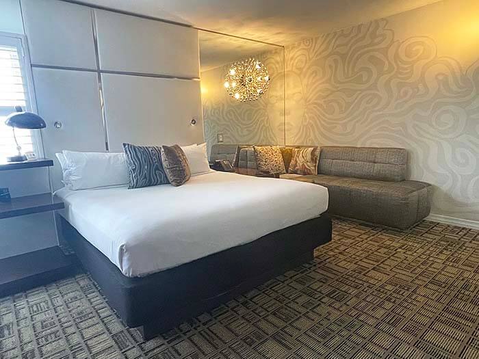 A king-size bed with white linens protrudes into the center of the room. Floor-to-ceiling mirrors and a subdued mural cover the walls.