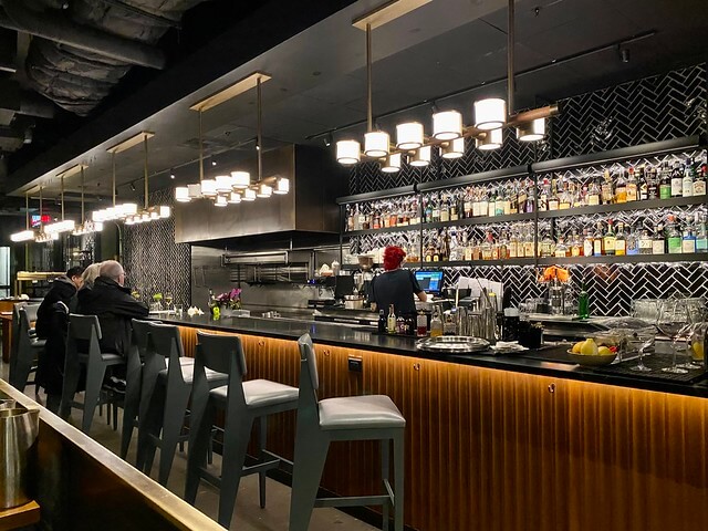 Hotel guests bellied up to the bar counter at Conversation Kitchen & Bar. A bartender with red hair prepares a drink with her back to the camera. 