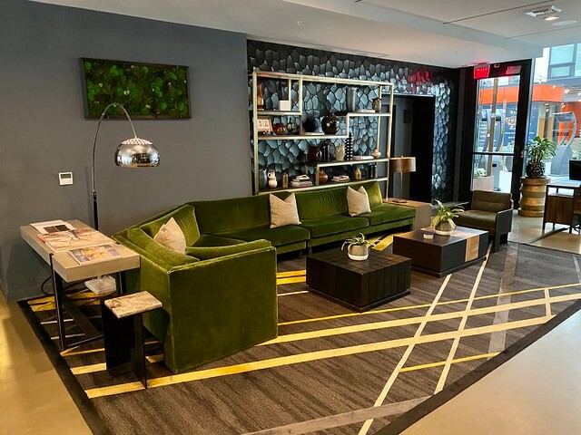 Large green velvet sofa sectional is the main focus of the hotel lobby. Shelves with sea glass on the wall make the lobby look like a cozy living room with plush pillows and succulent potted plants on 2 square wood tables. 