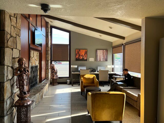 Best Western Sunridge Inn lobby with 2 yellow velvet low rise chairs by gas fireplace with rock wall. A hotel guest is seated on the sofa with his laptop, working. 