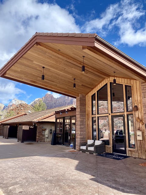 Wood portico covers the entrance to Cliffrose Springdale. Glass windows reflect red rocks across the street. Giant sandstone rocks loom behind the Springdale, Utah hotel entrance. 