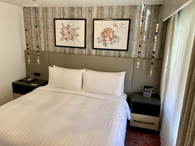 Cliffrose river side king bed with white duvet and four white pillows. Above the bed are two rose framed artwork pieces. A light gray, faux wood wall is behind the beige headboard that runs the length of the room. Wood, beaded blocks suspend on a rope on either side of the bed. Built in night stand hold a hotel telephone on one side and clock radio on the other side of the bed. 