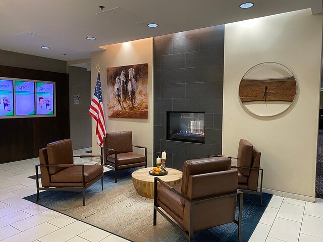 The small lobby area with gas fireplace, beyond reception desk at Holiday Inn Express Boise. Four brown leather chairs surround a small wood table. A gas fireplace is next to the chairs. An American flag is on a floor pole. Artwork of white horses running is on the wall. 