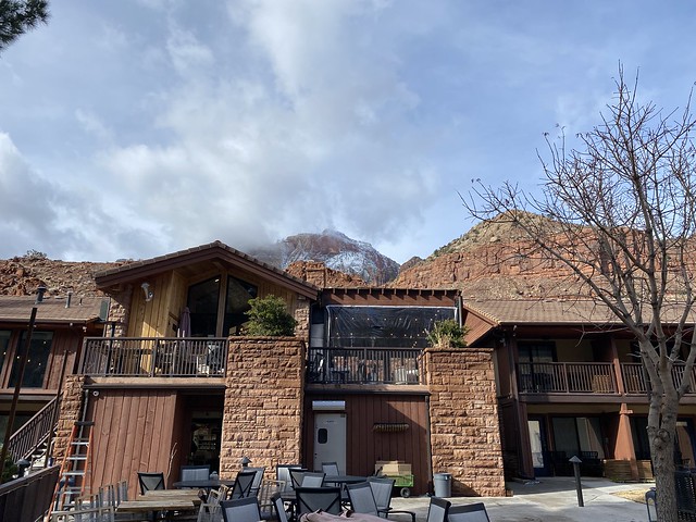 Exterior of Cliffrose Springdale, looking back at the hotel lobby and restaurant. Patio chairs and tables are below the hotel. Snow-dusted mountain peaks rise behind the Springdale, Utah hotel. 