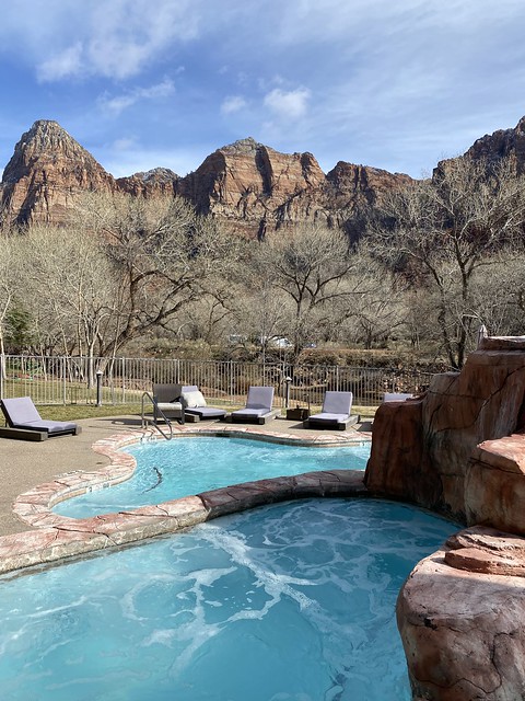 Outdoor hot tub, surrounded by 5 chaise lounges. Trees without leaves and rock formation are behind the hot tub at Cliffrose Springdale in Utah. 