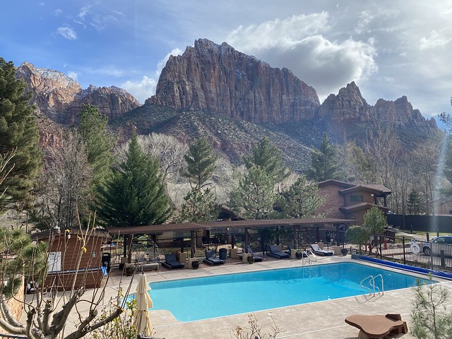 Cliffrose Springdale outdoor swimming pool. Four chase lounges are in a covered wooden cabana. Pine trees surround the back of the pool. Beyond, Watchman rock formation looms behind the hotel property in Southern Utah.