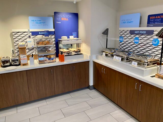 Holiday Inn Express hot breakfast buffet. Two connecting counters offering toast, pastries, cinnamon rolls. Adjoining counter holds hot breakfast entrees such as eggs and biscuits with gravy. 