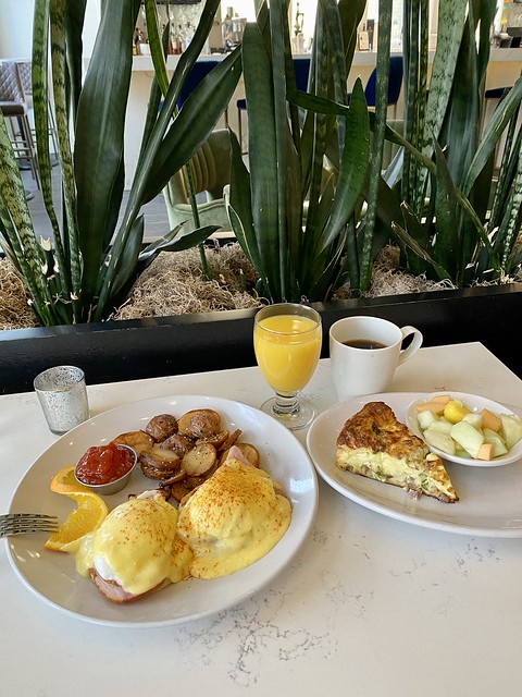 Eggs Benedict on 2 English muffins with hollandaise sauce, roasted potatoes. Second plate is leek and ham quiche with glass of orange juice and coffee. 