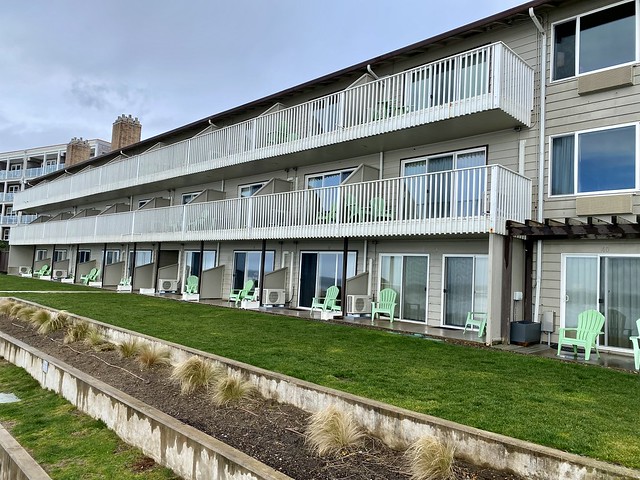 Three-story Coho Oceanfront Lodge from the back. Second and third floor hotel rooms have balconies, first floor rooms have small patio with stretch of green lawn. A tiered cement wall features dune grass on several rows. 