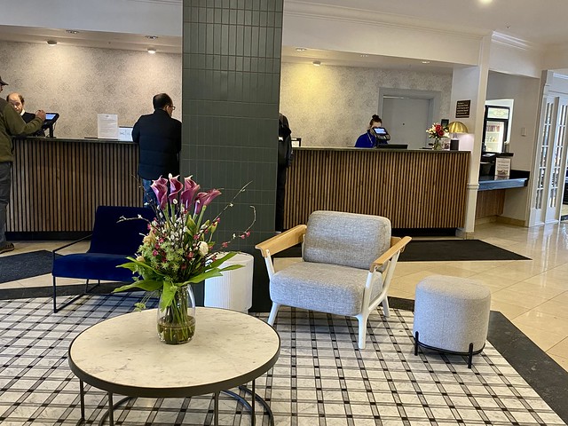 Registration area with hotel guests checking in at the counter. Two Crowne Plaza Portland hotel staffers assist guests behind the counter. Floral bouquet on circular table by reception.