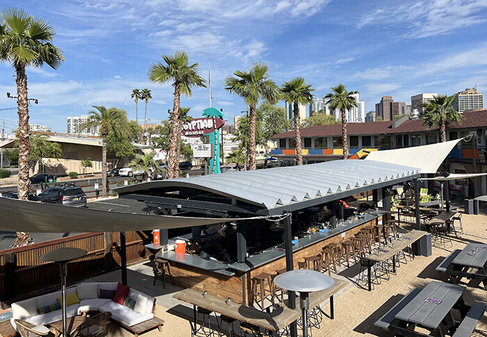 Outdoor bar with barstools and picnic tables in sand, surrounded by palm trees and blue sky.