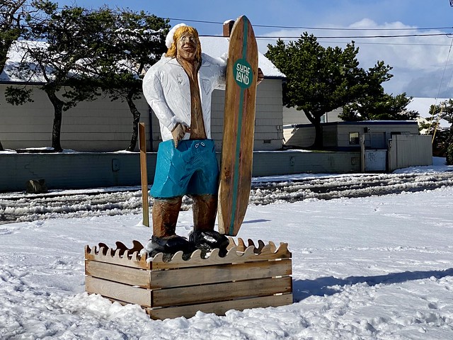 Wooden carved statue of a surfer dude, holding a wooden surfboard that says Surfland. He has on a white shirt and blue swim trunks, made of wood and painted. He is on a raised, wooden platform. A fresh blanked of snow surrounds the surfer guy wood carving. 