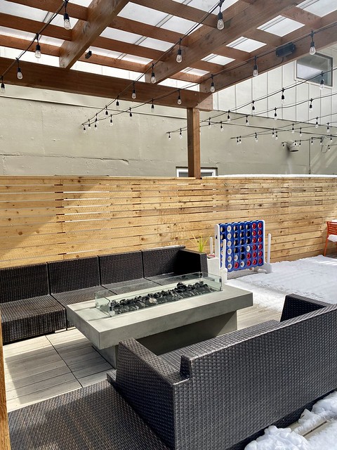 Outdoor firepit with gray wicker, bench-style sofas on either side of fire pit. a wood wall is behind the fire pit, separating the patio from the hotel. 