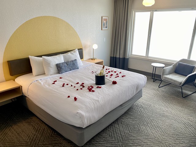 King bed in studio hotel room with red rose petals scattered on white bedspread. Ice bucket with bottle of white wine & 2 champagne flutes are in ice bucket. 