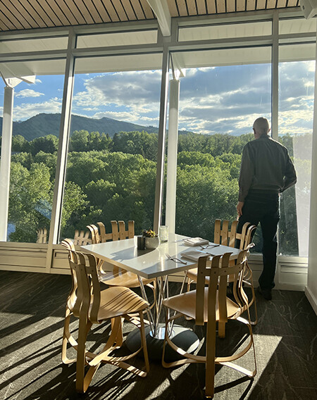 Sun shining into a restaurant with floor to ceiling windows and mountain view with man looking out.