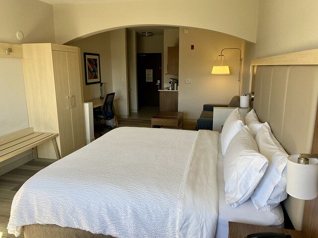 White bedding on Holiday Inn Express king bed. Wooden luggage rack and beige wood closet at the foot of the king bed. Small desk with chair, sleeper sofa and small wood table are beyond the king bed. A Small wet bar is next to the hotel room door, which is beyond the bed. 