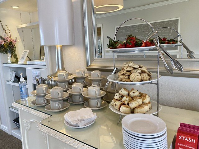 Scones, cookies and strawberries are displayed on a 3-tired tray, for hotel guests to enjoy, along with tea cups and saucers on the glass-topped buffet table.