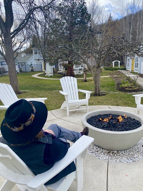 The author, Nancy Brown, sits in a white Adirondack chair by the circular gas fire pit. Two additional white Adirondack chairs sit vacant across from the lighted fire at Lithia Springs Resort. 