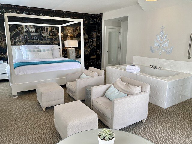 Water Lily suite with four-poster bed, two white chairs and white foot stools. A white soaking tub is to the right of the hotel room.