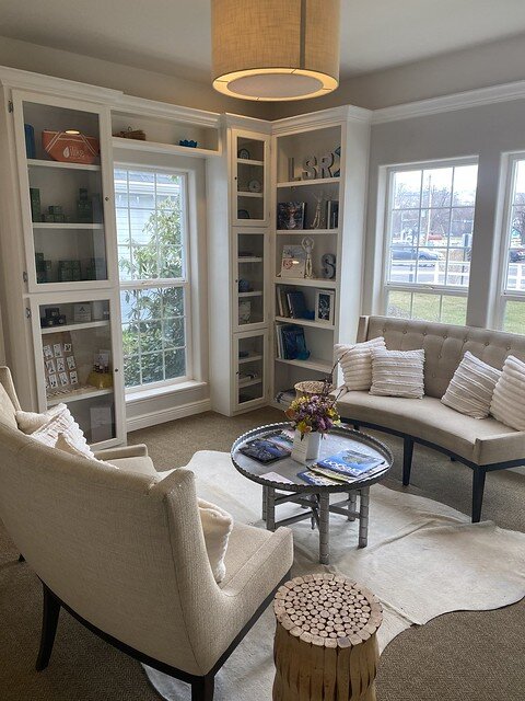 Waterstone Spa waiting room with 2 glass, floor to ceiling shelving units. Two white sofas are opposite a circular, small metal table with a fresh flower arrangement. Several magazines are on either side of the flower arrangement. 