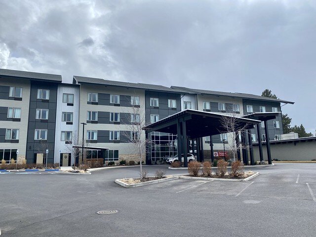 Best Western Premier Peppertree Inn Bend exterior. Four story hotel with covered portico entrance. A white SUV is parked under the portico. The large, concrete parking lot in front of the hotel sits empty.