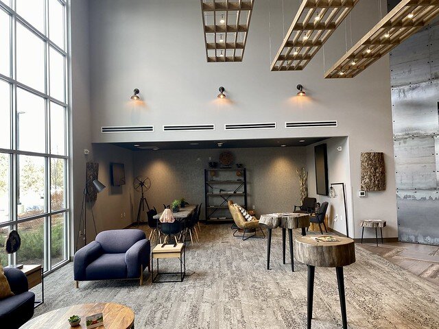 BW Peppertree hotel lobby with wall of glass windows at the hotel entrance. The lobby has a beige, multi-colored area rug, several circular wood tree slab small tables for gathering and a large, blue overstuffed chair to the left of the tables. At the rear of the lobby is a wooden rectangular tables that seats 8 chairs. 