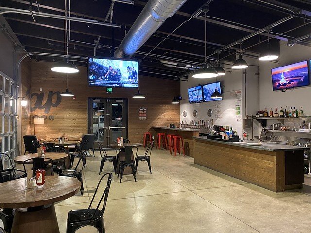 BW Peppertree Pub & Eatery with 4, circular wood tables. Two and three metal, black chairs surround each table. A bar and pay station is to the right of the pub restaurant. Beyond the bar is a bank of chairs and a granite countertop for solo and couple dining inside the restaurant. 