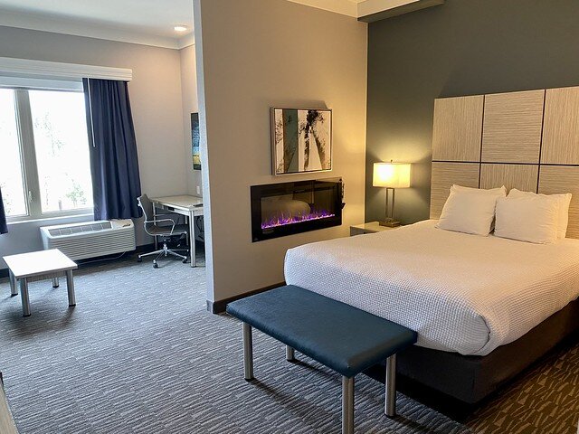 Queen bed, white bedspread. Blue cloth and metal bench at base of bed. Gas, rectangular fireplace with small artwork of abstract tree trunk on wall above gas fireplace.