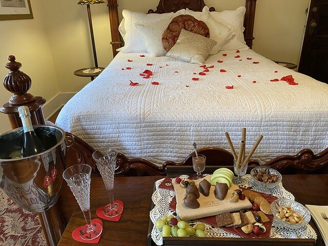 Lady Florence queen bed with white bedspread, covered in cloth rose petals. At the based of the bed is a wooden hope chest with a chocolate charcuterie board and all sorts of chocolate goodies, including 4 chocolate covered strawberries. To the left of the wooden board is an ice bucket stand with bottle of chilled Prosecco. Two cut crystal glasses are set on the hope chest, to the right of the ice bucket. 