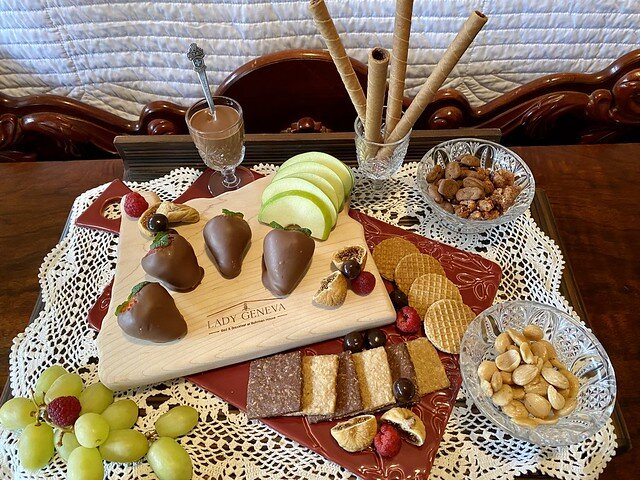 Chocolate-covered strawberries, sliced green apple, small carafe of melted chocolate to drizzle on apple pieces, crackers and fresh grape cluster & raspberries are scattered on the Lady Geneva logo wood cutting board. Guests are able to keep the board.