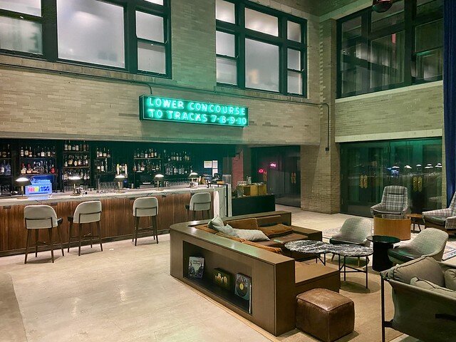 Several bar chairs are in front of the hotel bar. Lots of 750 ml liquor bottles are on bar shelves. Neon green sign reads Lower Concourse To Tracks 7-8-9-10. Hotel lobby l-shaped brown leather sofa and 2 olive green chairs separate bar from living room area. 