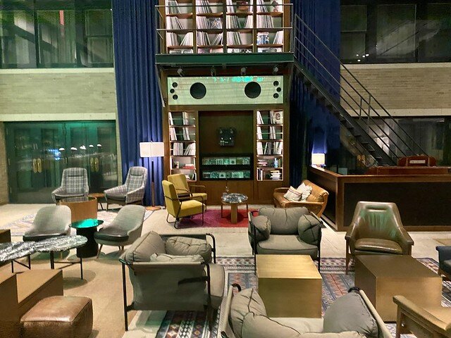 Two-story music room with 2 Eggleston Works speakers on back wall. Collections of records displayed on wall shelves above the speakers. Stairs climb to the record display. Lounge chairs are arranged in the hotel living room to the right of the hotel bar.