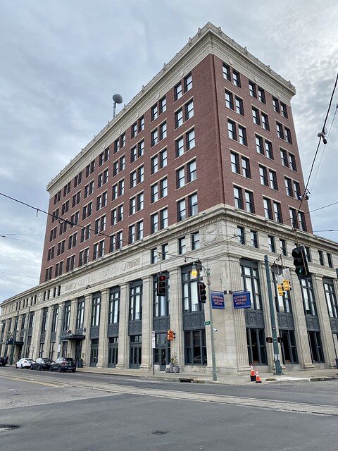 Exterior brick facade of the 8-story Central Station Memphis hotel. Black wire holds directional street lights above street intersection, in front of hotel. Three cars are parked on the street, in front of the hotel. 