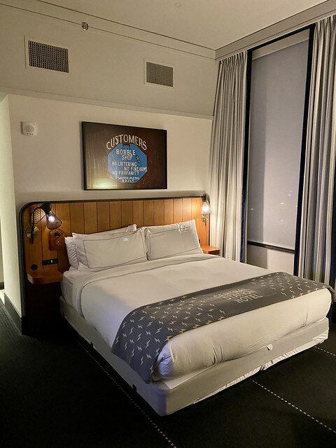 Hotel king bed with gray blanket on base of bed. The bed has white bedspread and several white pillows with a wood headboard. Above the headboard is an antique sign saying Customers Bobble Shop, No Loitering, No Firearms, No Profanity. Watch metal rails.