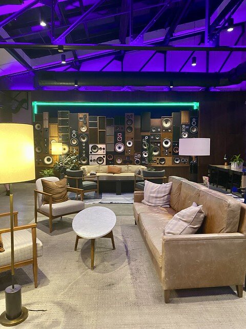 Hotel lobby with floor to ceiling stacked stereo speakers. A long beige leather sofa is in front of the speakers. A light beige leather sofa and 2 wood chairs are by the oval table. Purple lights iluminate the ceiling and metal rafters of the hotel. 